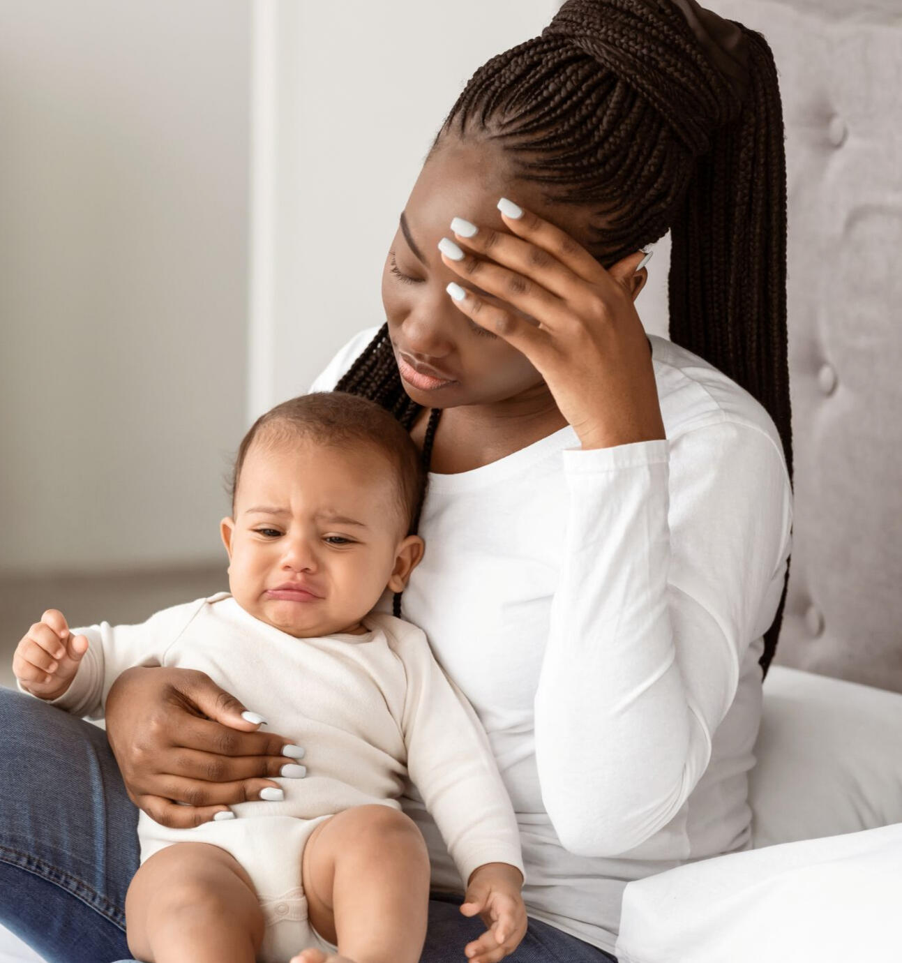 Mother sitting on a bed holding her baby while looking tired and overwhelmed during a difficult moment after childbirth